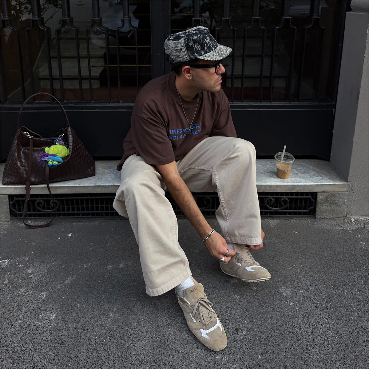 A man sits on a stone bench in front of a black metal gate, wearing a brown t-shirt, beige wide-leg pants, and beige sneakers. A brown leather handbag sits to his left, and an iced coffee cup is on the bench to his right.
