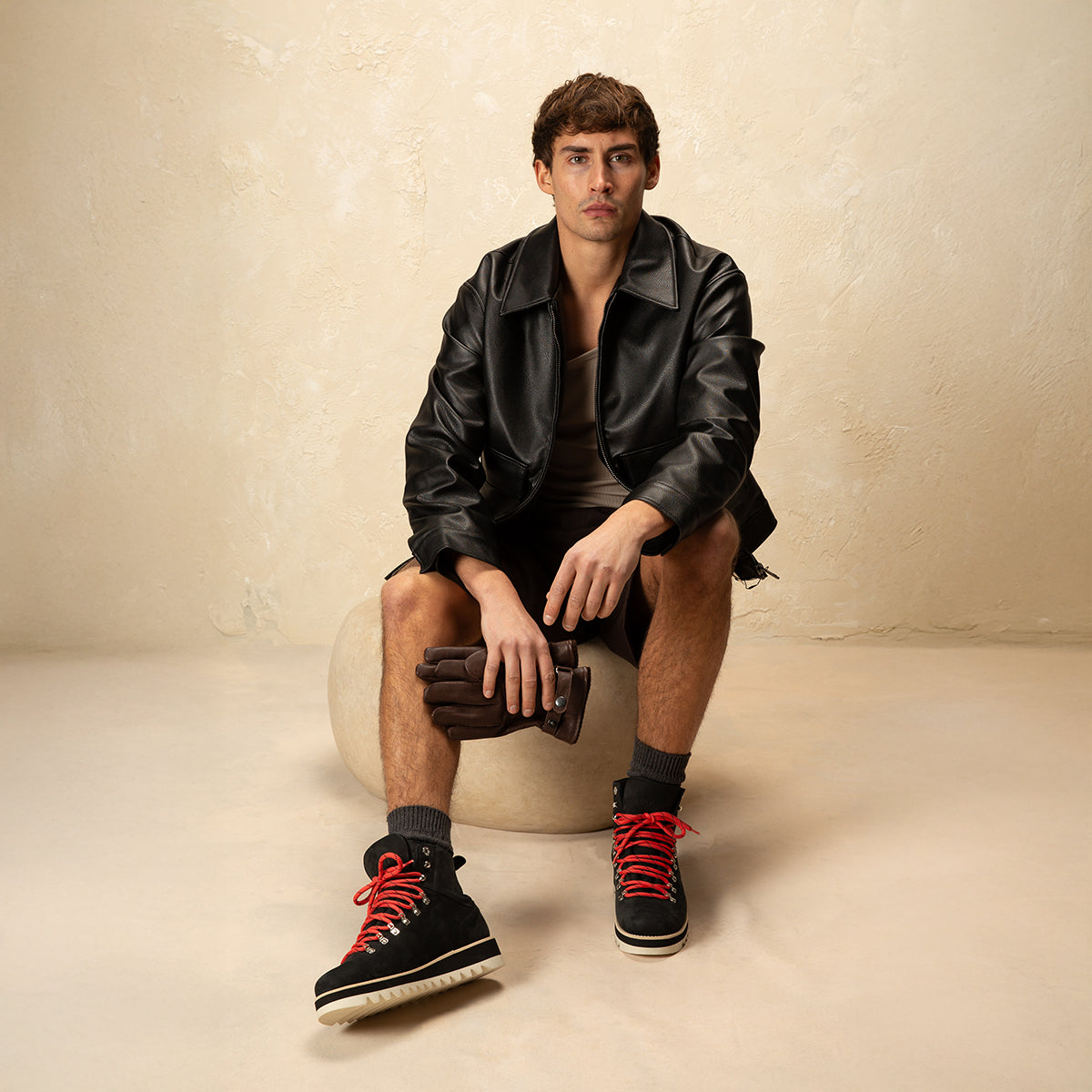 Male model sitting on a stool and wearing black leather jacket, shorts, black boots with red laces