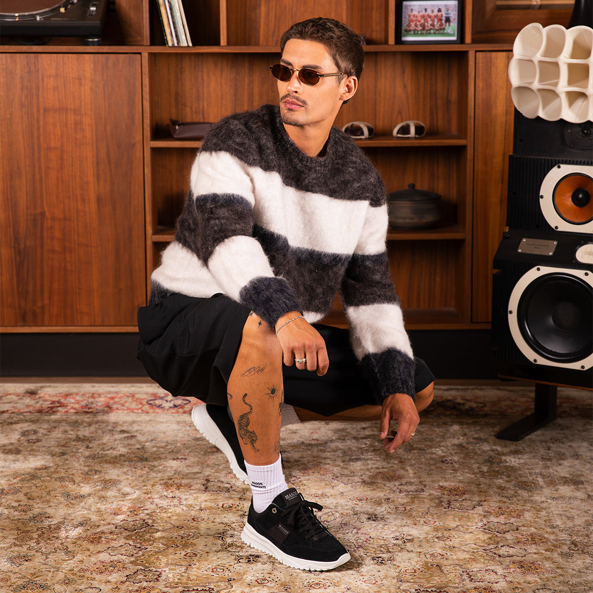 A man wearing a fuzzy black and white striped sweater, black shorts, white socks, and black sneakers crouches in a room with wooden shelves and a speaker.