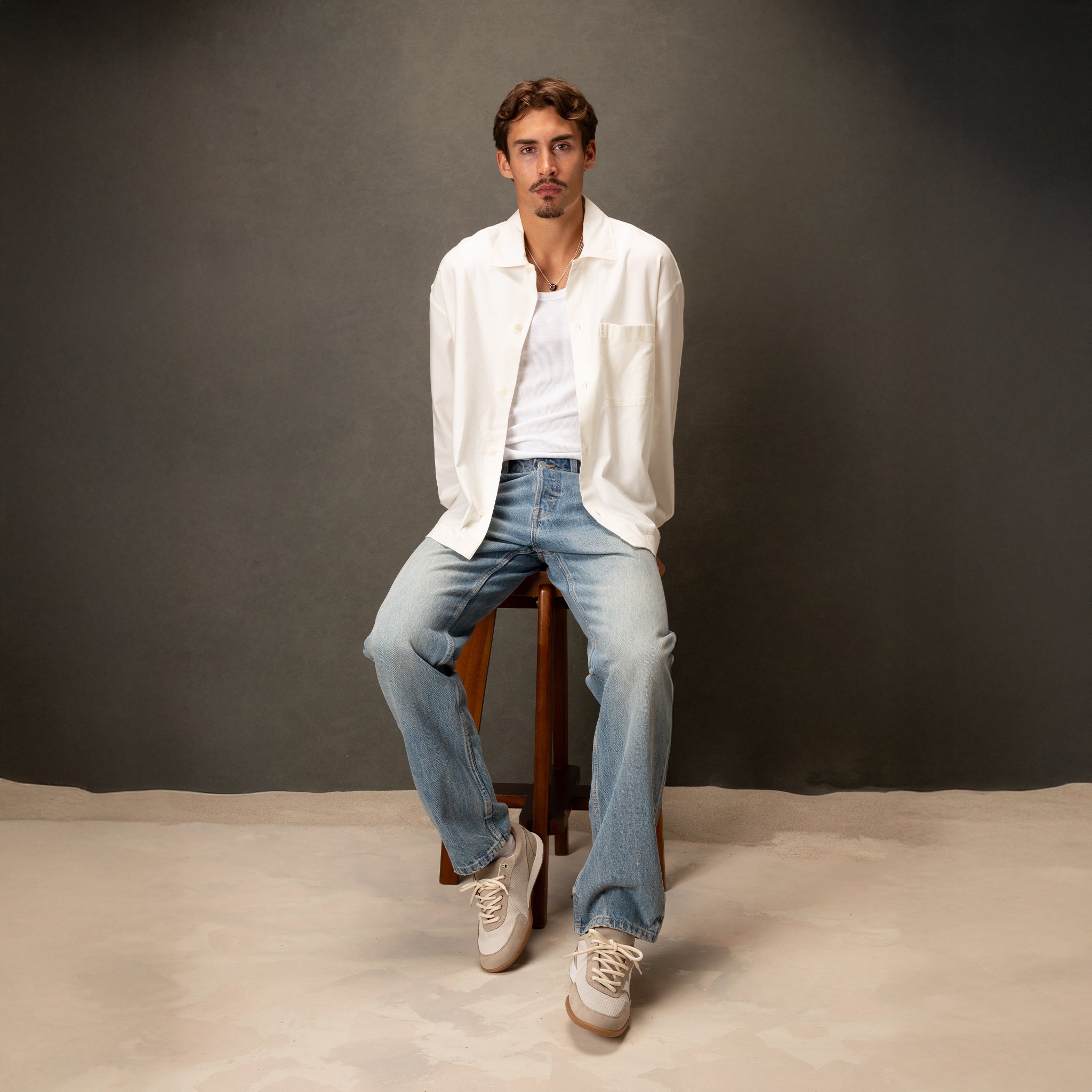 Man sitting on a stool wearing a white shirt, blue jeans, and white sneakers against a gray background