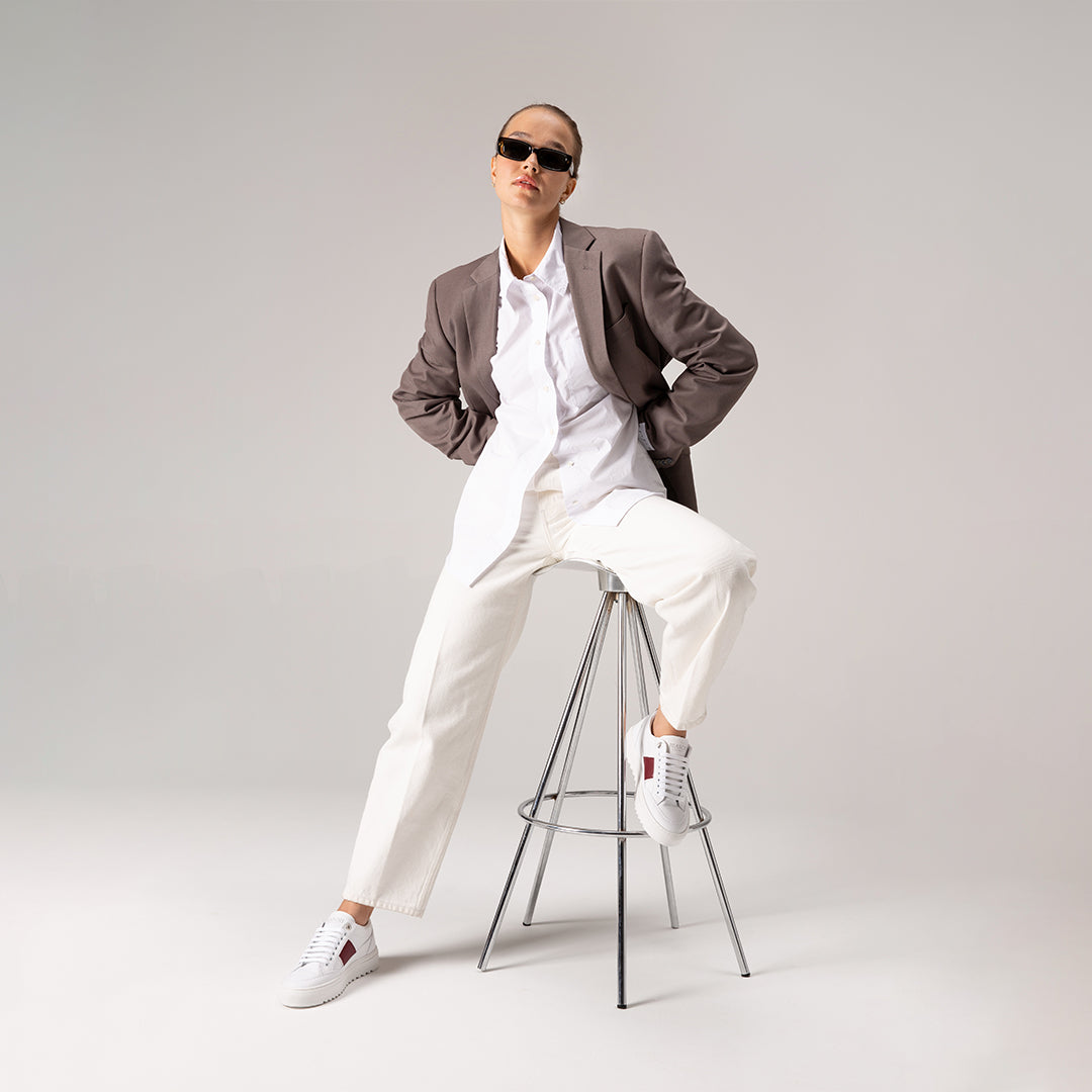 Female model wearing a gray blazer over a white outfit with white sneakers, sitting on a stool against a plain background