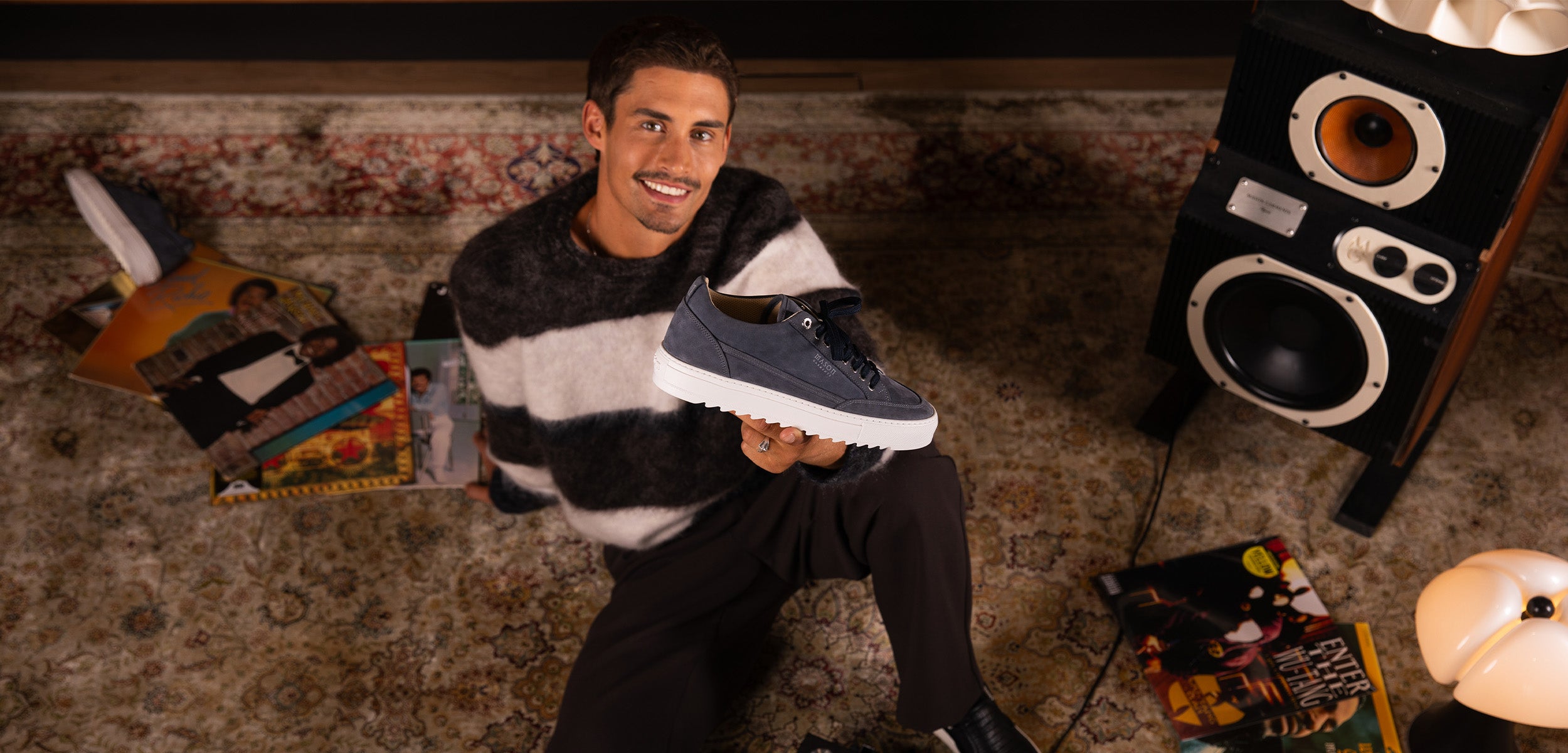 A man in a black and white striped sweater sits on a patterned carpet, holding up a dark blue sneaker with a white sole. Vinyl records and a large speaker are scattered around him in a cozy, retro-styled room.