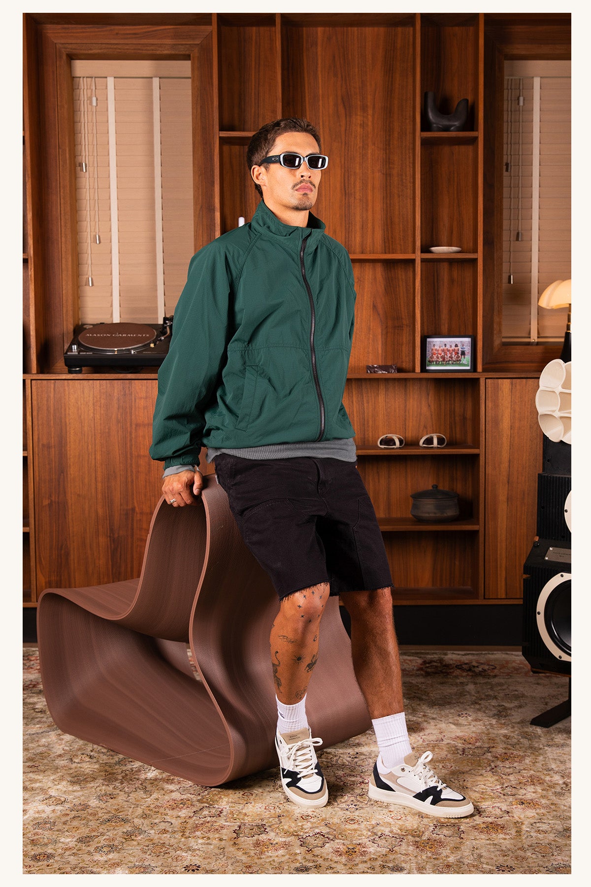 A male outfit with a dark green zip-up jacket, black shorts, white socks, and black-and-white Mason Garments sneakers, in a room with wood paneling, holding a brown sculptural chair.