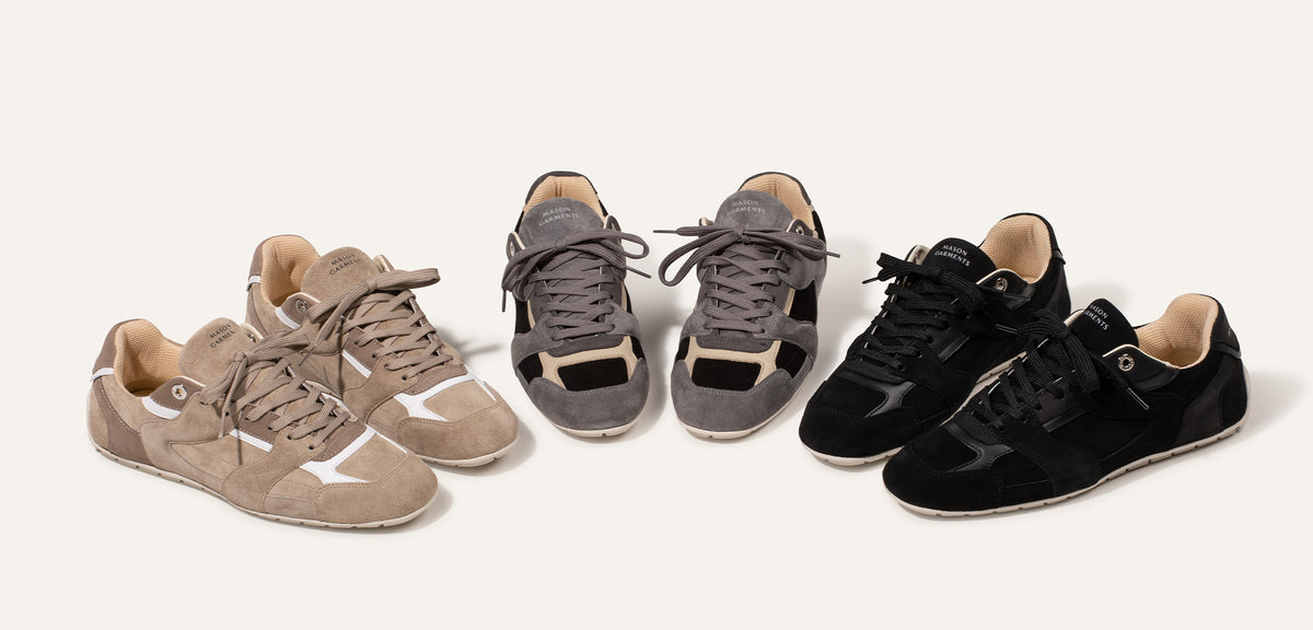 Three pairs of suede sneakers arranged on a light beige background, shown in beige, gray, and black colorways. The shoes feature a classic low-top design with suede panels, matching laces, and subtle white contrast accents.