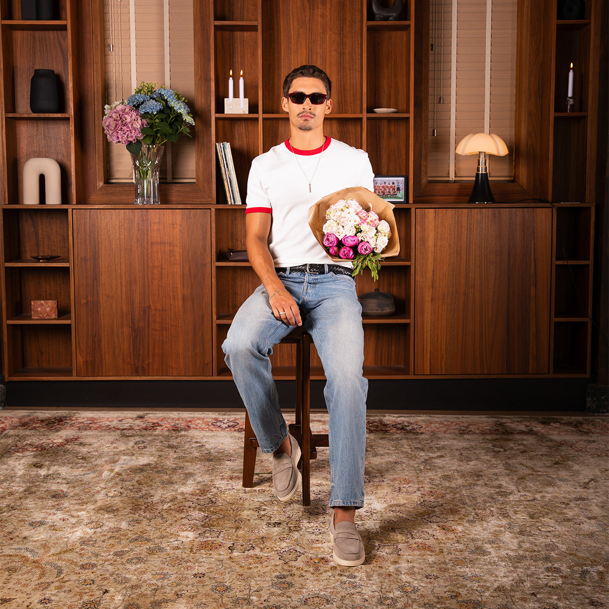 A man wearing a white t-shirt with red trim, blue jeans, beige penny loafers and sunglasses sits on a wooden stool in a room with a wooden bookshelf background, holding a bouquet of pink and white flowers.