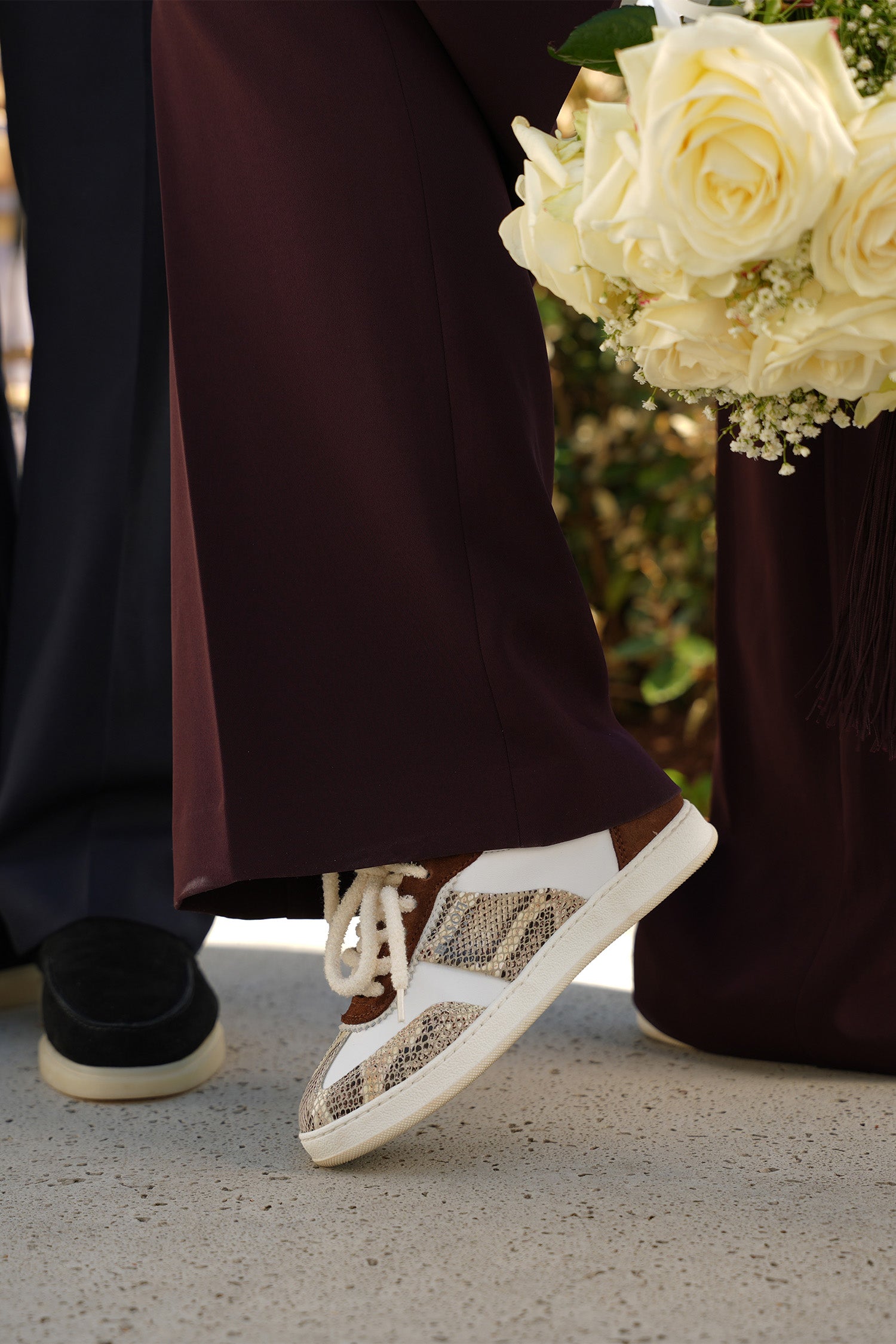 Person wearing burgundy pants with white and brown sneakers and holding a bouquet of white roses