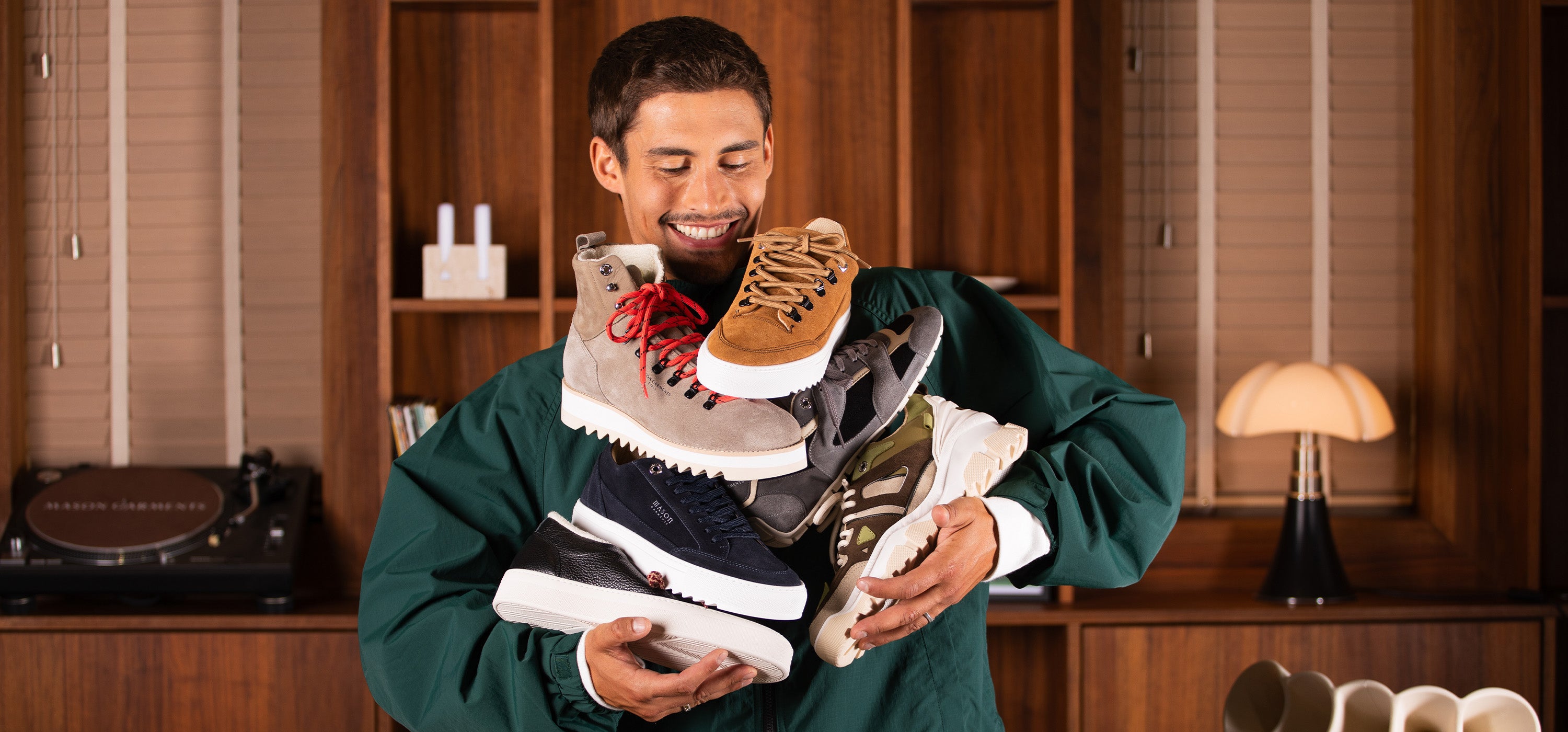 A man in a green sweatshirt holds a large stack of various Mason Garments sneakers in front of a warm wood-paneled room background.