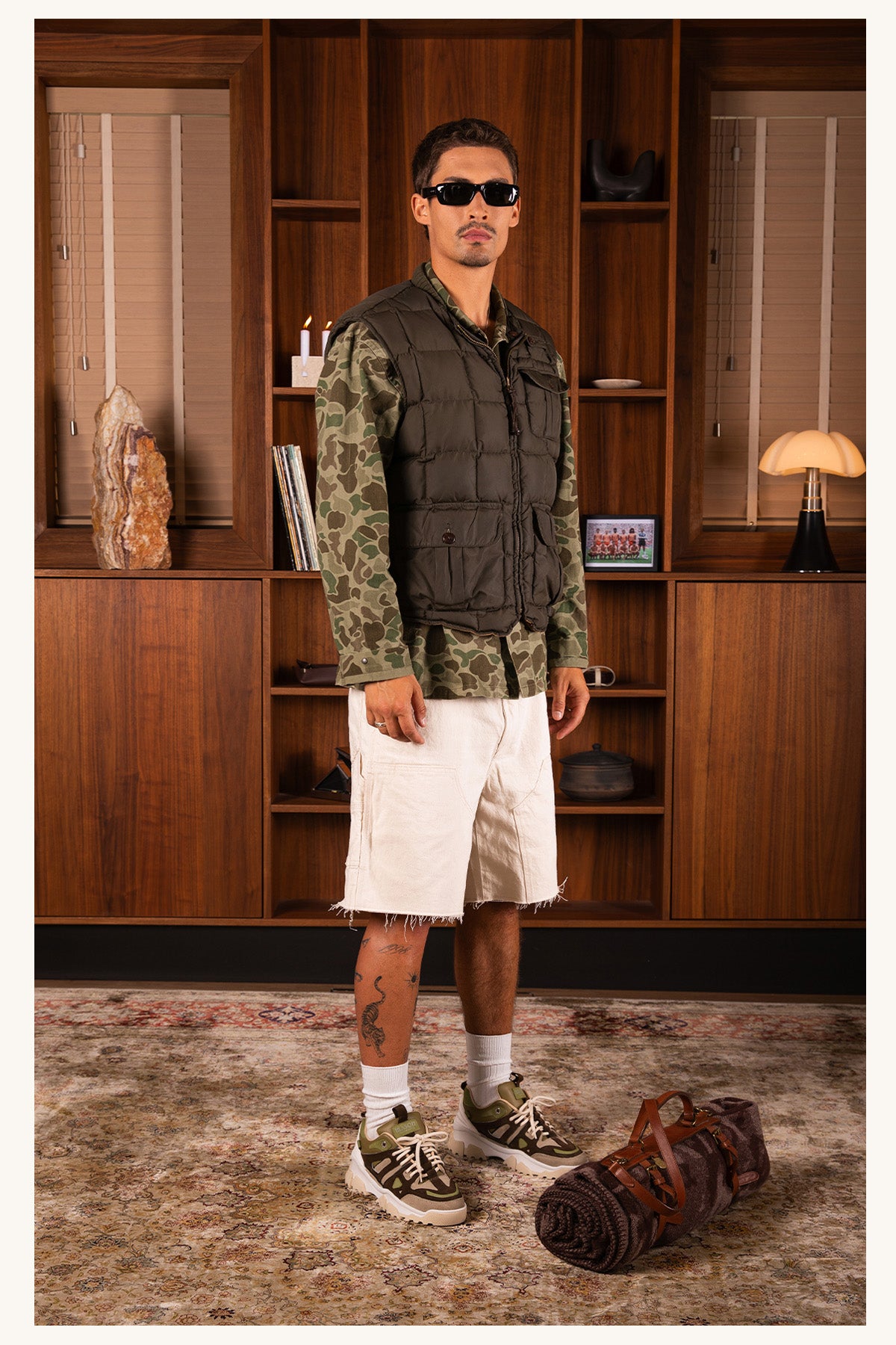A man wearing a dark olive green quilted puffer vest over a camouflage long-sleeve shirt, paired with off-white shorts, white socks, and olive green Mason Garments sneakers, standing in front of a wooden bookshelf.
