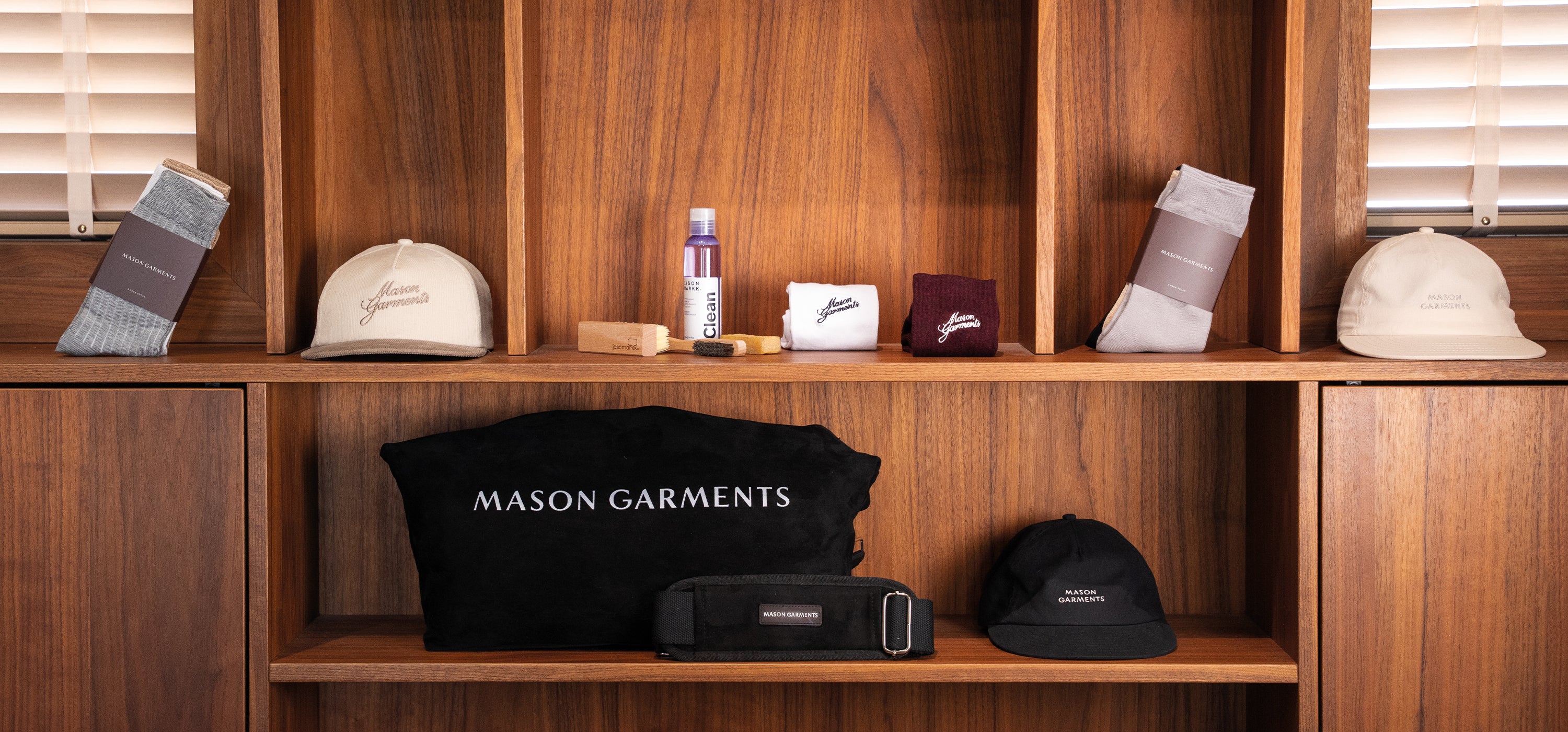 A wooden display shelf showcasing Mason Garments products, including a black branded tote bag, a black cap, a white cap, socks and cleaning products.