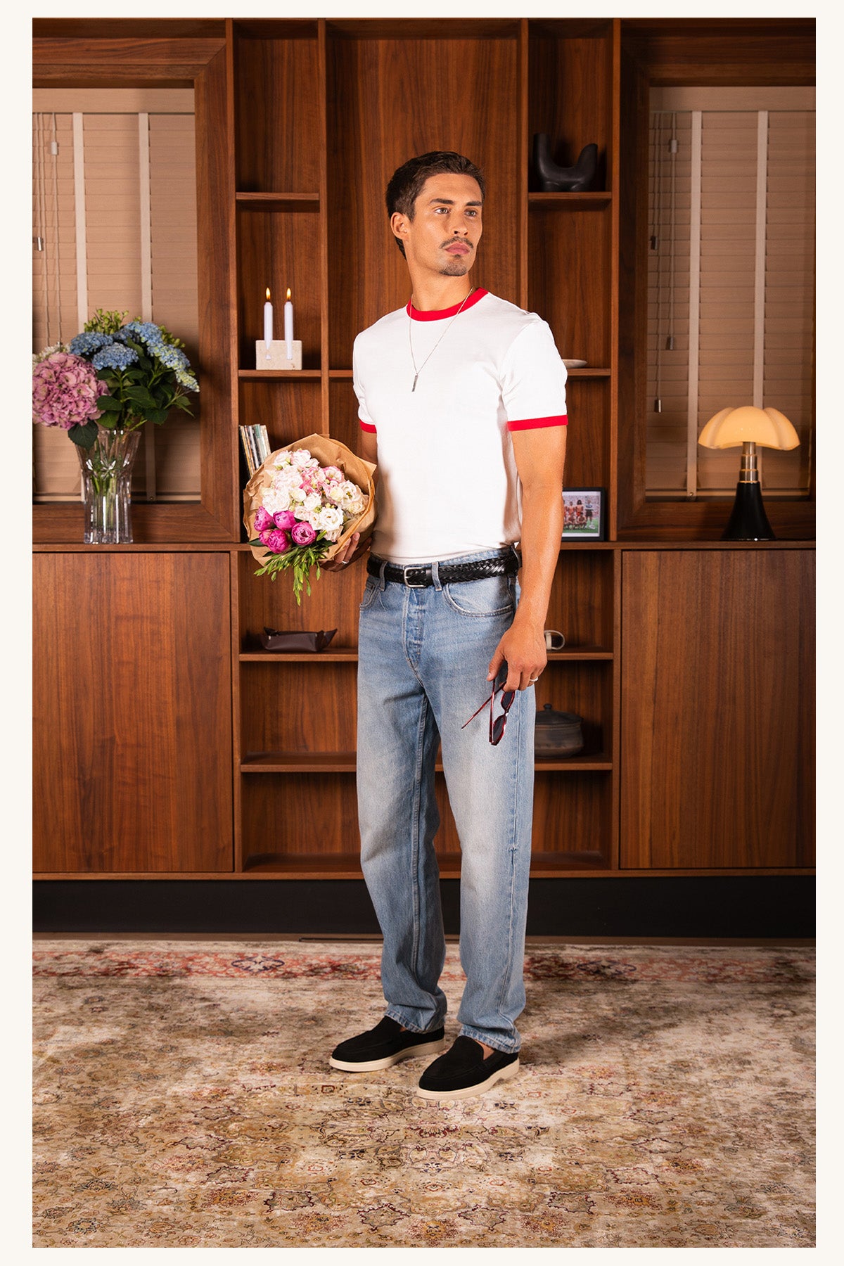 A man wearing a white short-sleeve t-shirt with red trim, light blue jeans, and black slip-on Mason Garments loafers stands in a room with wooden bookshelves, holding a bouquet of flowers.