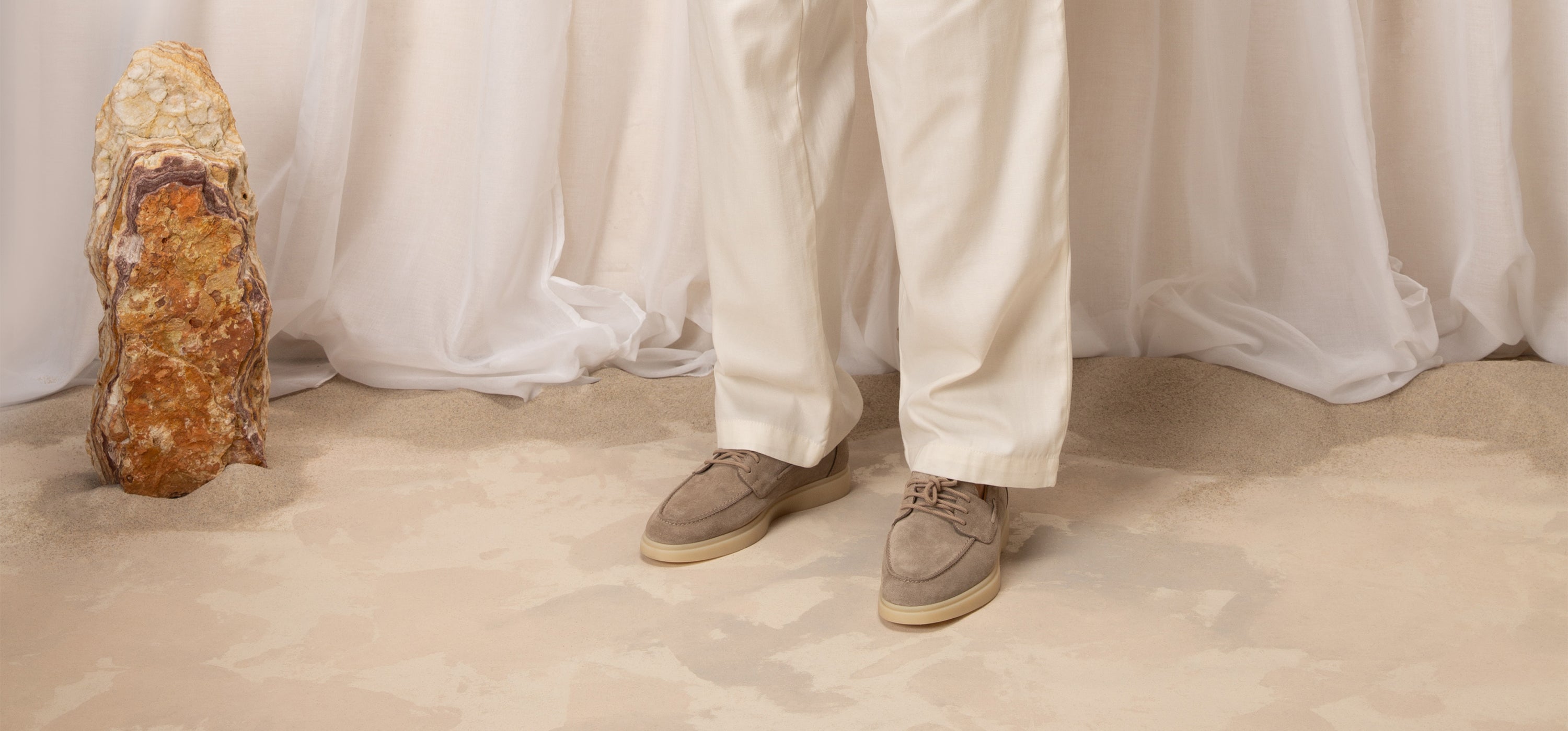 Close-up of a person's feet wearing brown shoes and white pants, standing on a sandy surface with a large rock in the foreground.