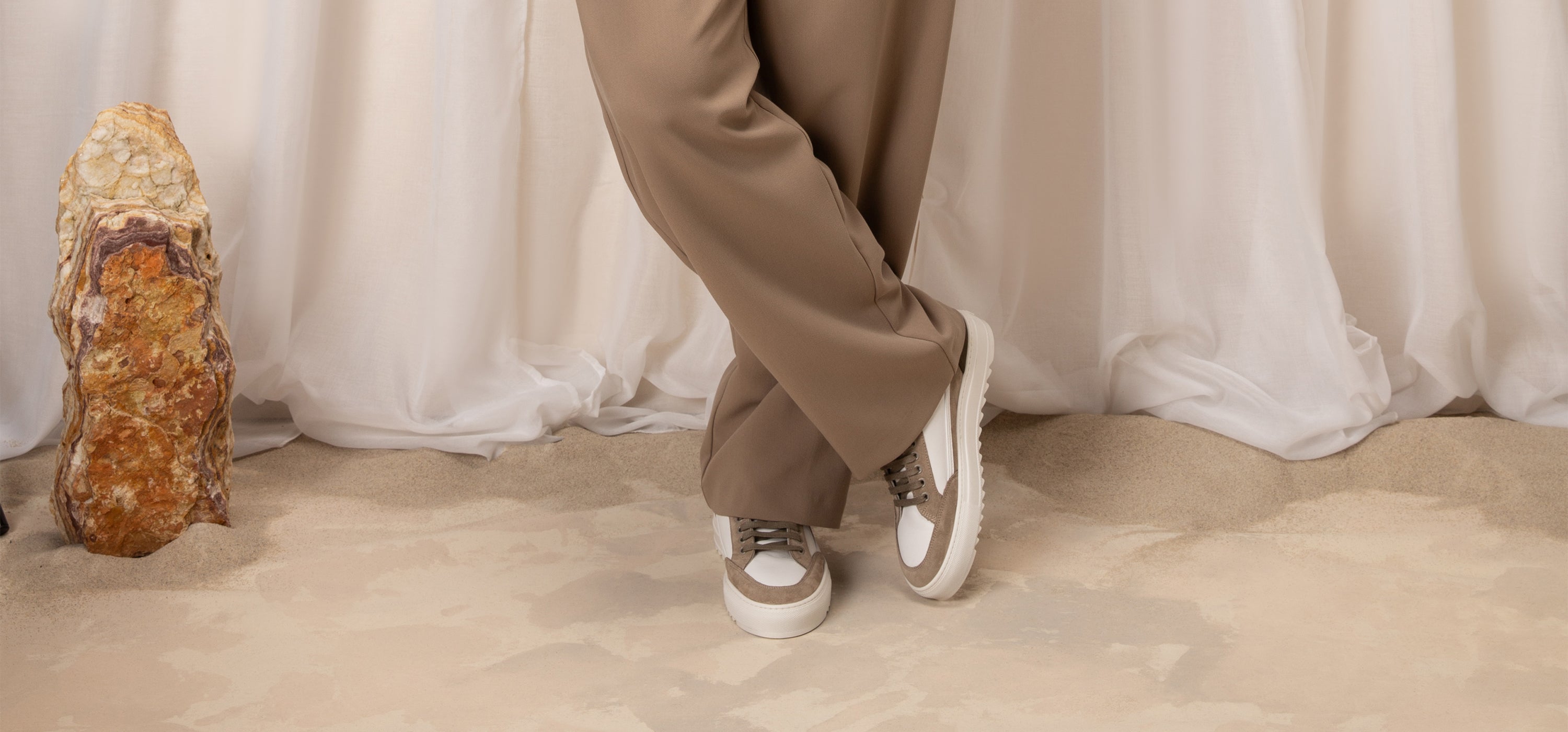 Person wearing beige pants and white sneakers standing on a beige carpet with a white curtain in the background.