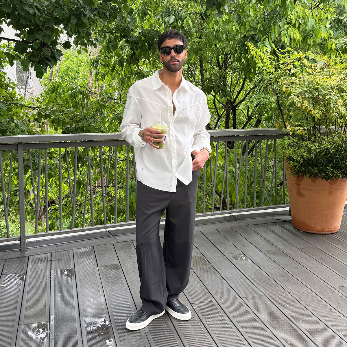 Man standing on a wooden deck holding a drink, surrounded by greenery in leather black slipons