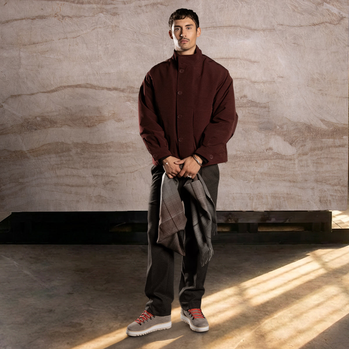 Man wearing a brown coat, dark pants and brown boots against a textured wall.