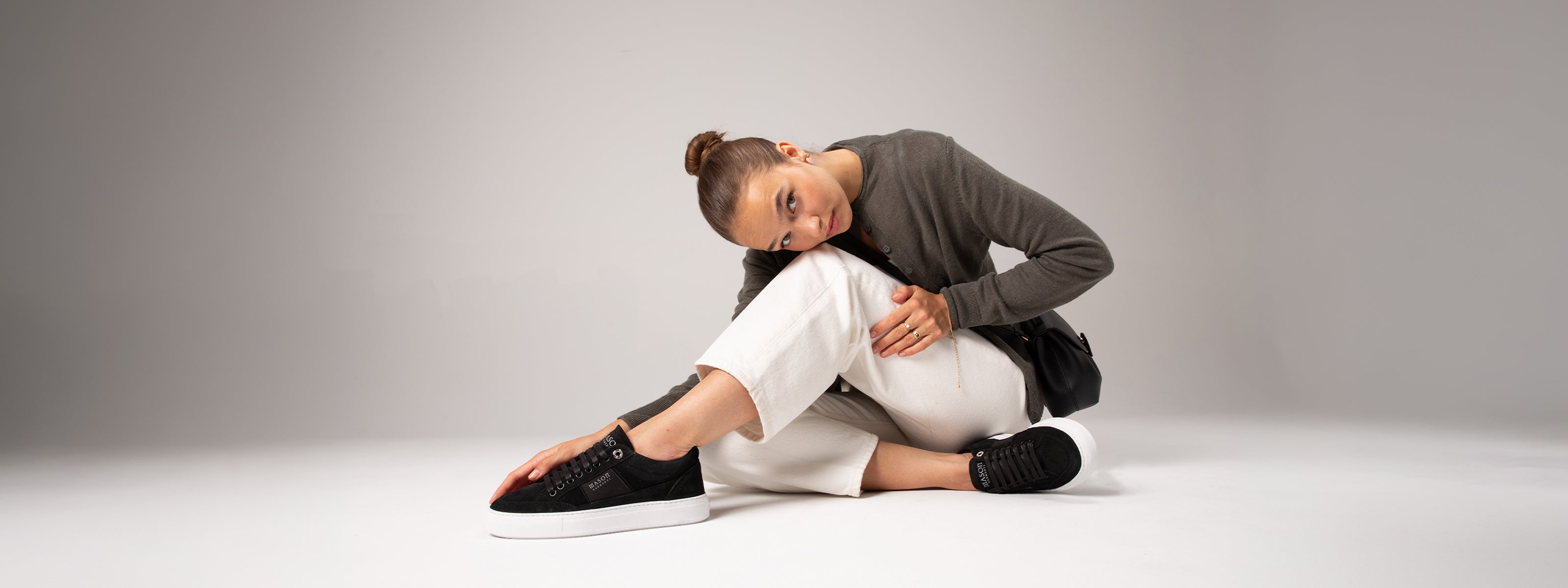 Woman sitting on the floor wearing black shoes and neutral outfit against a plain background