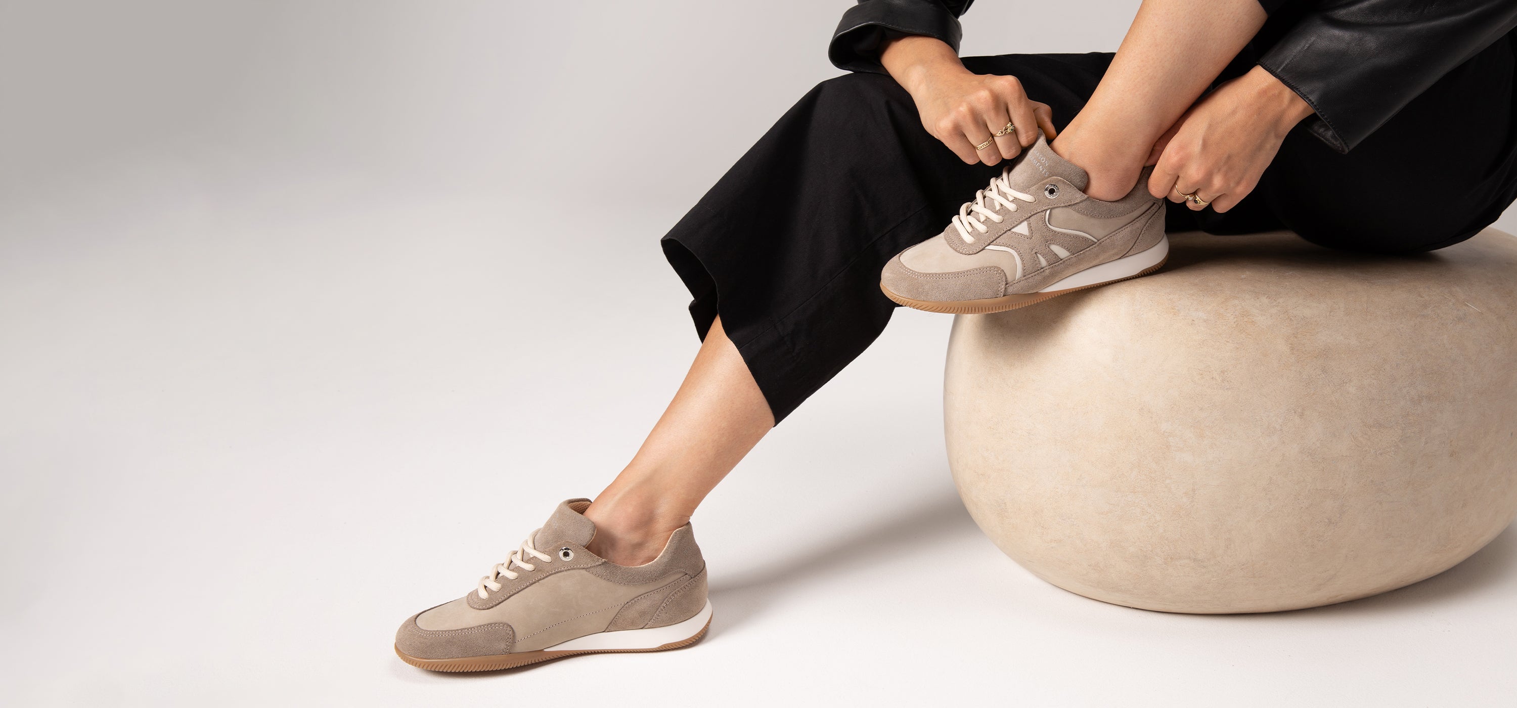 Person wearing beige sneakers sitting on a white stool against a light gray background